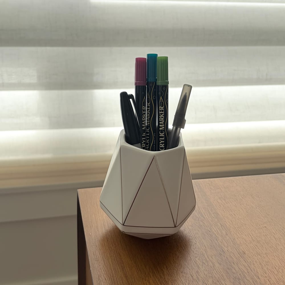 A white hexagonal ceramic pen cup with thin gold lines, holding three colored acrylic markers and two pens on a wooden desk by a window