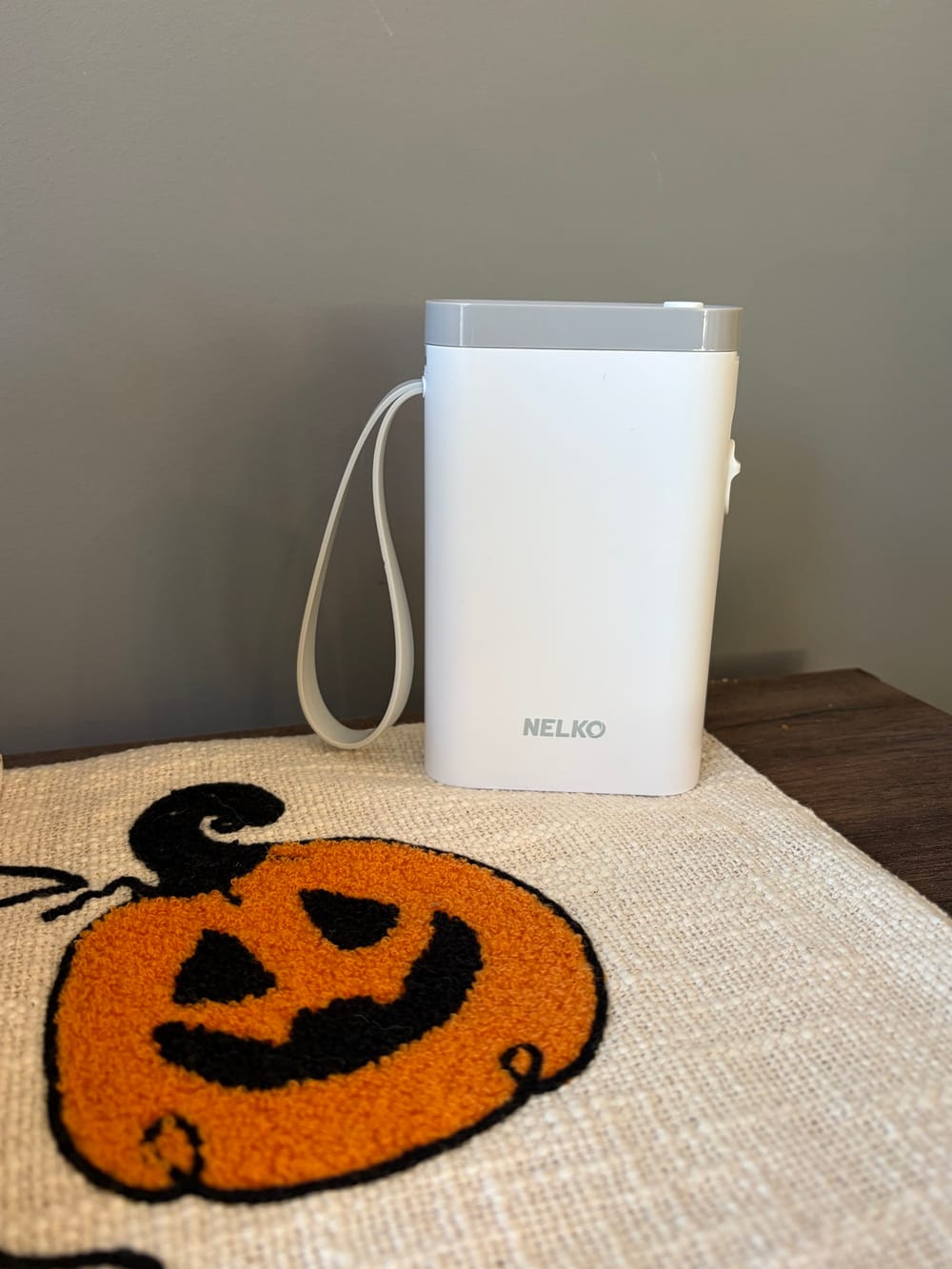 A white Nelko P21 label maker with a gray wrist strap, standing on a table runner embroidered with a jack-o'-lantern